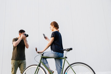 Profile of fashionable photographer taking a photo of smiling beautiful model using a mobile phone while sitting on bike. Outdoors.