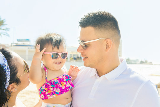 Young man father holds his little cute baby toddler daughter girl on hands young mother woman smiling looking at each other. Sunny summer day oceanfront. Family love togetherness unity concept