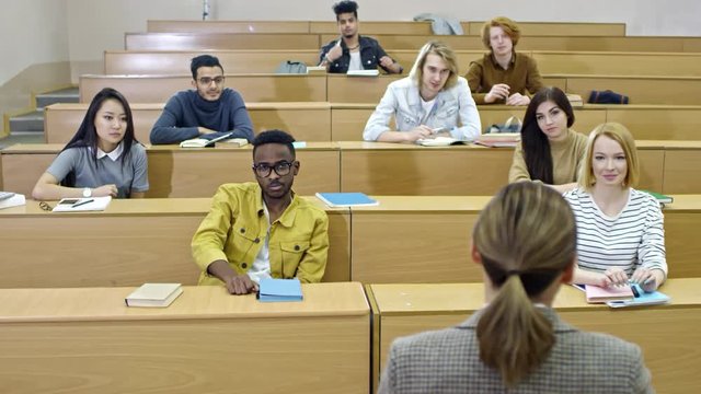 Group of young multi ethnic students sitting in lecture hall and listening to female professor seen from her back, one of them raising hand to ask question