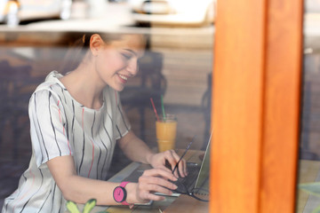 Young freelancer with laptop working in cafe, view through window