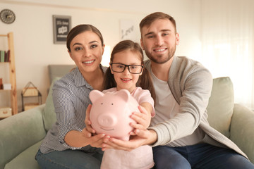 Happy little girl with her parents sitting on sofa and holding piggy bank indoors. Money savings...
