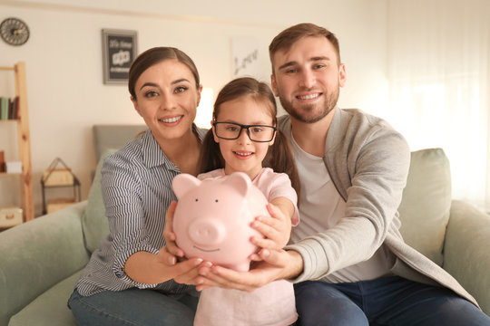 Happy little girl with her parents sitting on sofa and holding piggy bank indoors. Money savings concept