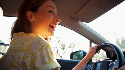 Medium shot of a young beautiful woman driving and smiling at someone in the passenger seat with a big smile and the sun shining on her face.