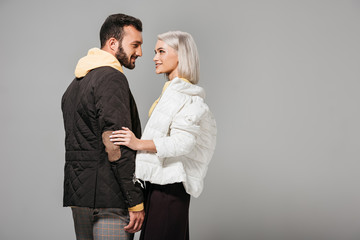 side view of stylish couple of models in autumn outfits looking at each other isolated on grey background