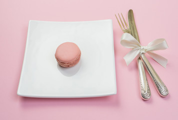 Silver fork and knife with white ribbon and plate with a maracon on pink background