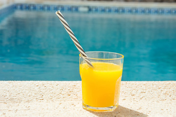 Glass of freshly pressed tropical fruits orange juice with striped straw standing on deck of swimming pool. Bright sunlight. Summer vacation relaxation travel luxury. Authentic atmosphere lifestyle