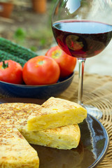 Traditional Spanish omelet tortilla frittata with potatoes eggs cut out wedge organic vegetables tomatoes cucumbers glass of red wine on rattan table in garden outdoors. Summer Day. Lifestyle