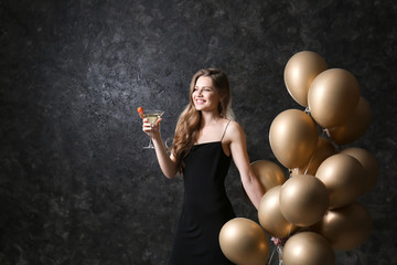 Beautiful young woman with balloons and cocktail on dark grey background