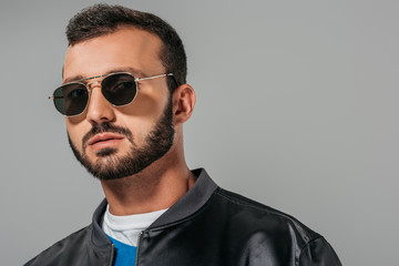 handsome man posing in stylish bomber and trendy sunglasses,  isolated on grey