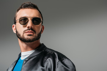 Obraz premium bearded man posing in black bomber and sunglasses, isolated on grey