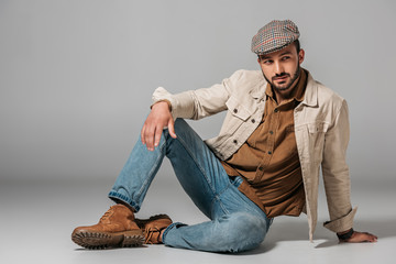 bearded man posing in corduroy shirt, jeans and autumn jacket and tweed cap, on grey