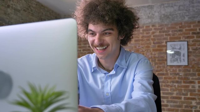 Happy businessman with curly large hair cheering about victory, winner sitting at table with laptop, modern office background