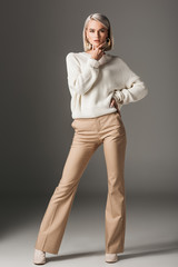 elegant fashionable woman posing in white sweater and beige pants, on grey