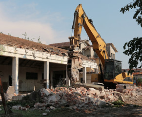 Obraz premium Excavator in front of a semi demolished building, among rubbles. The demolition is done for an urban redevelopment of the area