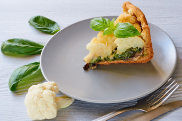 A piece of cauliflower pie on the gray plate decorated with fresh basil leaves and vintage silver knife and fork. Vegetarian healthy cauliflower tart on the gray kitchen background. Close up view