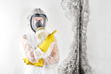 A professional disinfector in overalls and a mask points a finger at the black mold on the wall....