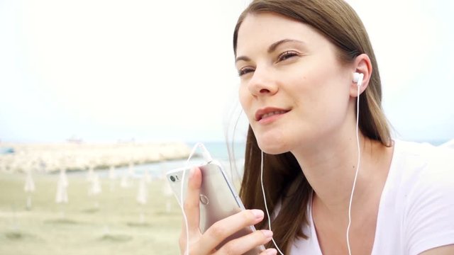 Happy smiling young woman using mobile on beach alone. Female listening music via cellphone in slow motion. Vacation during off-season on Mediterranean Sea