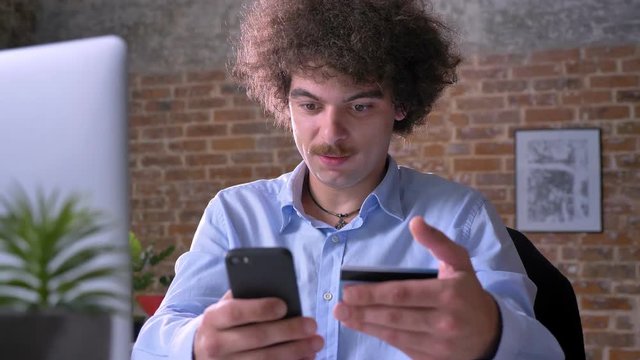 Funny nerdy office worker shopping on phone and paying with credit card, sitting at table in modern building