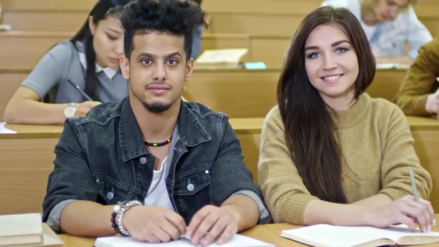 Tracking shot of two young students of different ethnicities sitting at desk in lecture hall, looking at camera and smiling