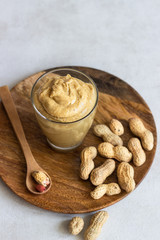 Creamy peanut butter in a glass jar and wooden spoon on light grey background. Copy space.
