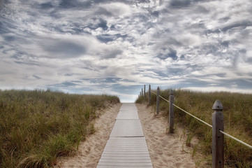 A wooden path leading among the sands and grass on the beach of the Antaltiechi coast. Road going up into the clouds. USA. Maine.
