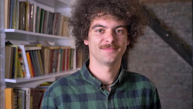 Funny nerdy man with curly hair and mustache standing in library and looking at camera, cheerful and positive