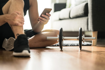 Fototapeta Man using smartphone during workout at home. Online personal trainer or on mobile phone. Internet fitness class or video course. Taking a break. Lazy guy with cellphone while training with dumbbell. obraz