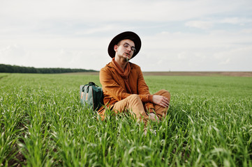 Stylish man in glasses, brown jacket and hat with bag posed on green field.