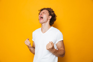 Photo of happy man 20s in casual t-shirt rejoicing and screaming with clenching fists, isolated...