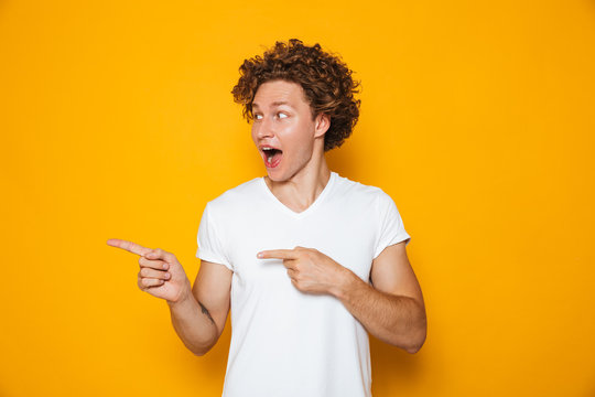 Excited cheerful man 20s with brown curly hair gesturing finger aside on copyspace, isolated over yellow background