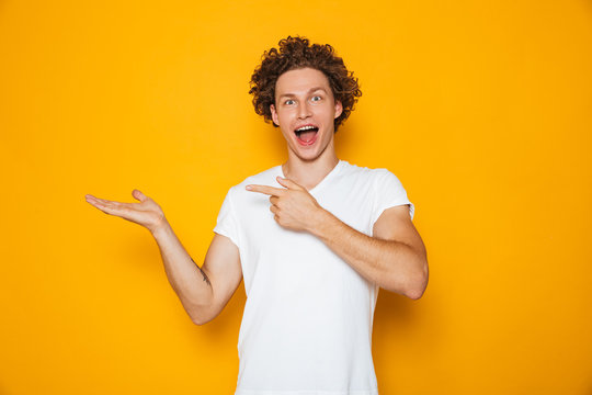 Young happy man 20s with brown curly hair gesturing finger aside on copyspace at palm, isolated over yellow background