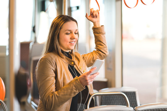 Woman Standing In Train, Tram Or Bus Holding The Handle And Using Mobile Phone. Happy Female Passenger Texting With Smartphone While Riding In Public Transportation.