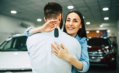 Happy young hugging couple chooses and buying a new car for the family in the dealership.