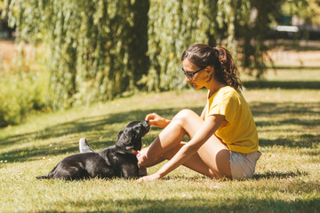 Naklejka na ściany i meble A young woman is sitting on the grass and playing (or feeding) with a dog / picture of a girl with sporty outfit having fun in a sunny garden