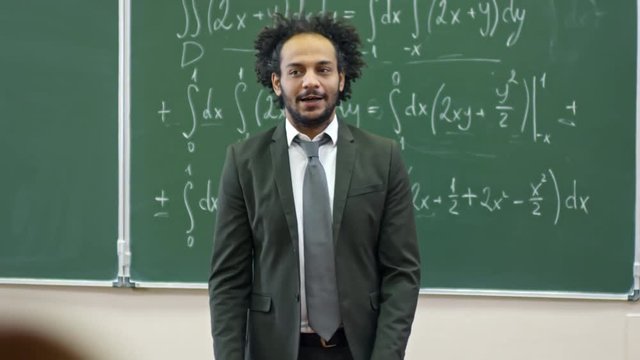 Medium shot of middle eastern man with curly hair standing at blackboard with equations written on it and explaining topic to students