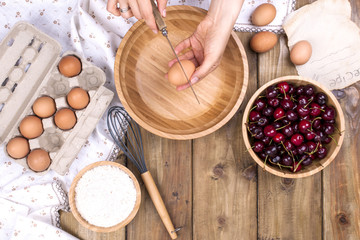 Female Hands break the egg into a wooden bowl. Preparation of dough for a pie with a cherry. Summer homemade pastries. Top view. Copy space.