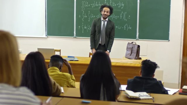 Tracking shot of friendly university professor leaning on chair when standing at blackboard and talking to students during lecture on mathematics