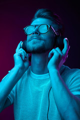 Handsome man in glasses enjoys listening to music with headphones. Neon studio portrait - 216136390