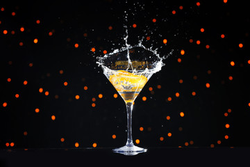 Glass with splashing cocktail and slice of orange against blurred lights