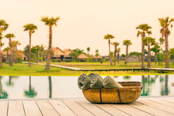 Naklejka premium Rolled bathing towels in a basket, on a wooden deck, next to an infinity pool reflecting palm trees and bungalows
