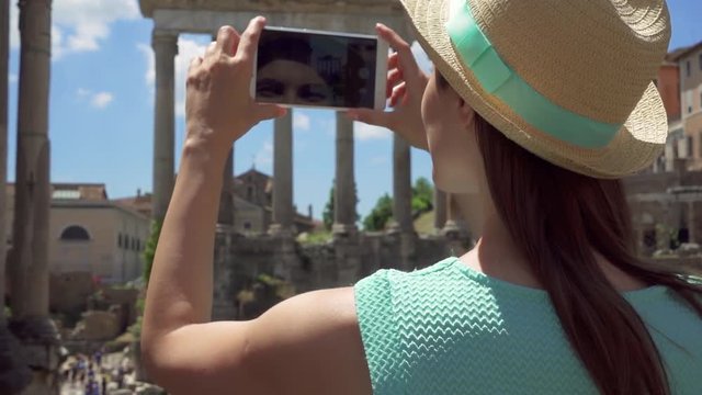 Woman in hat near ancient ruins Forum Romanum taking photo on mobile phone in slow motion. Happy female tourist taking picture of Roman forum in center of Rome, Italy. Student travel through Europe