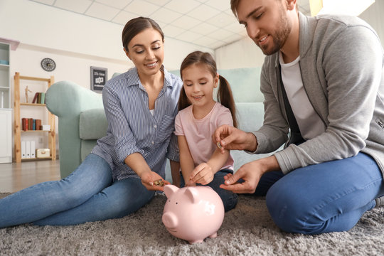 Happy little girl with her parents sitting on carpet and putting coin into piggy bank indoors. Money savings concept