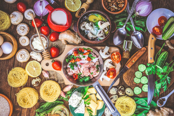 chicken meat in a frying pan with vegetables and mushrooms in batter with egg, herbs and milk. different food ingredients on a wooden background.