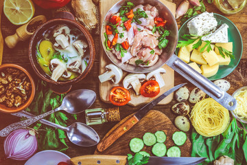 chicken meat in a frying pan with vegetables and mushrooms in batter with egg, herbs and milk. different food ingredients on a wooden background.