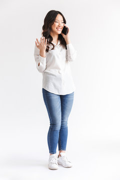 Full length photo of joyous chinese woman with long dark hair holding and talking on mobile phone, isolated over white background in studio