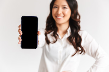 Blurry photo of smiling asian woman with long dark hair standing and showing smartphone at camera, isolated over white background in studio