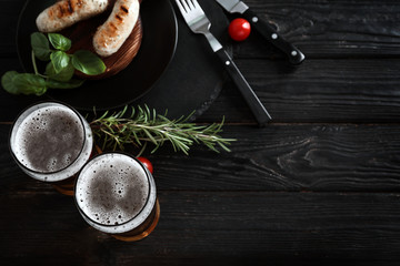 Glasses of delicious beer with grilled sausages and herbs on wooden table