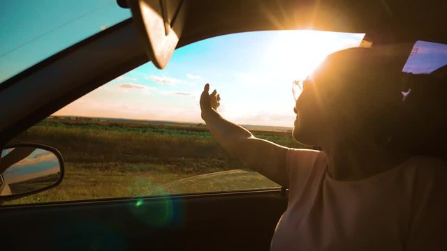 girl waving her hand in the window sunlight the wind In car slow motion video. Young happy young girl drives a car a holds her hand out from the window. Road trip lifestyle, travel and freedom concept