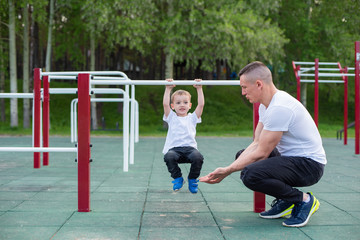 A strong athletic father insures the boy on sports equipment and teaches him to pull himself up on the horizontal bar in the summer park. Strong athletic father teaches his son to catch up on the bar.