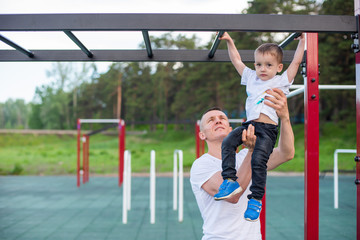 Obraz premium A strong athletic father insures the boy on sports equipment and teaches him to pull himself up on the horizontal bar in the summer park. Strong athletic father teaches his son to catch up on the bar.